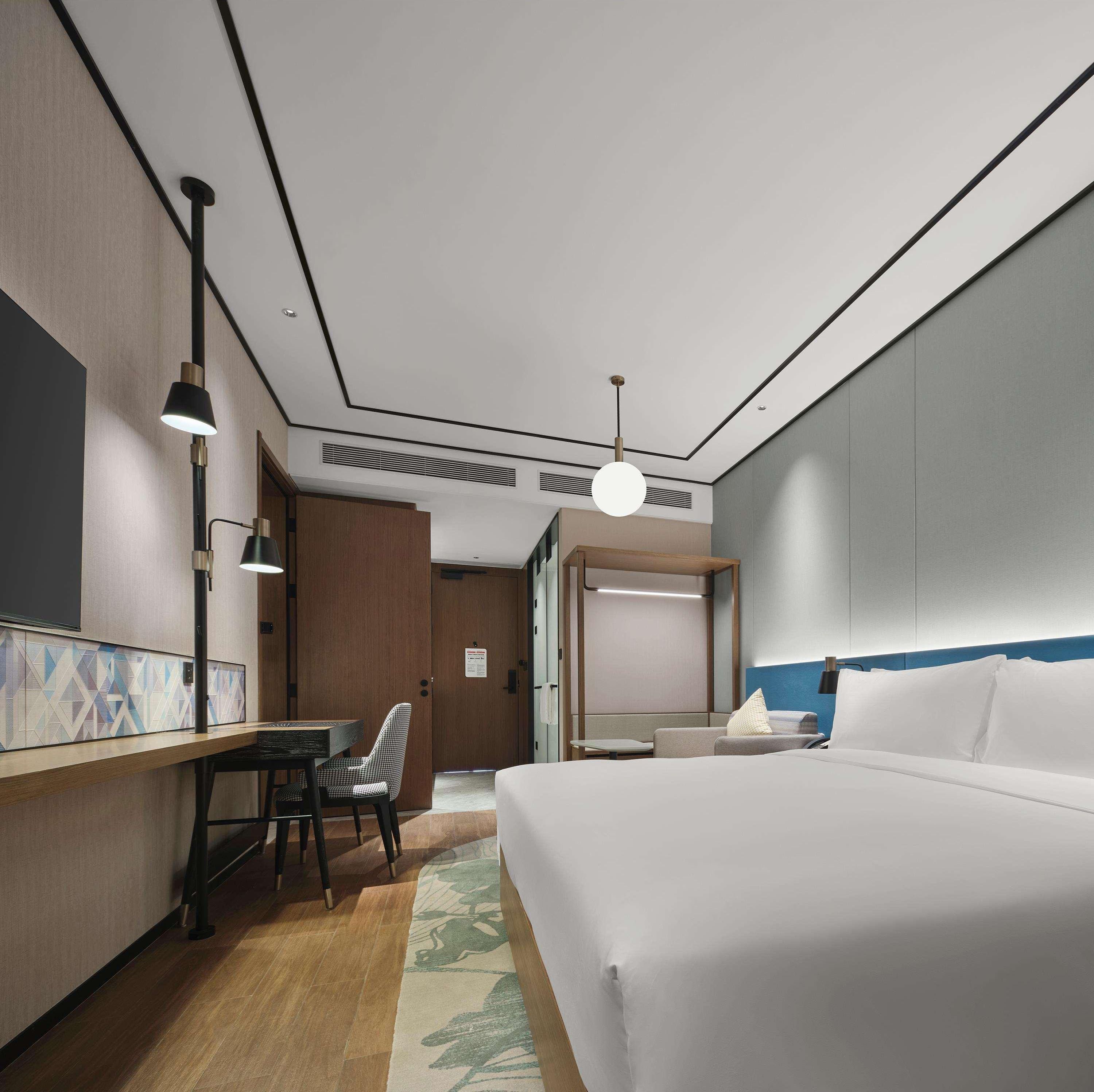 A hotel room with a bed, a desk with a chair, a hanging light fixture, and a door leading to another room.