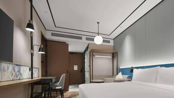 Una habitación de hotel con una cama, un escritorio con silla, una lámpara colgante y una puerta que da a otra habitación.