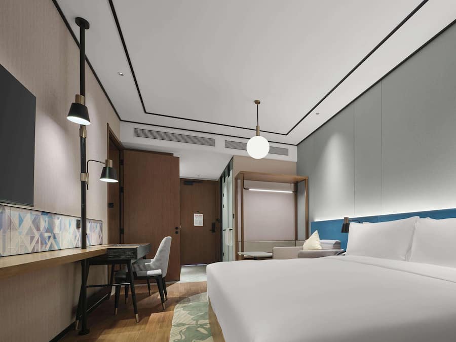 A hotel room with a bed, a desk with a chair, a hanging light fixture, and a door leading to another room.