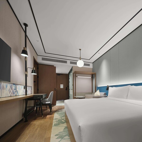 A hotel room with a bed, a desk with a chair, a hanging light fixture, and a door leading to another room.