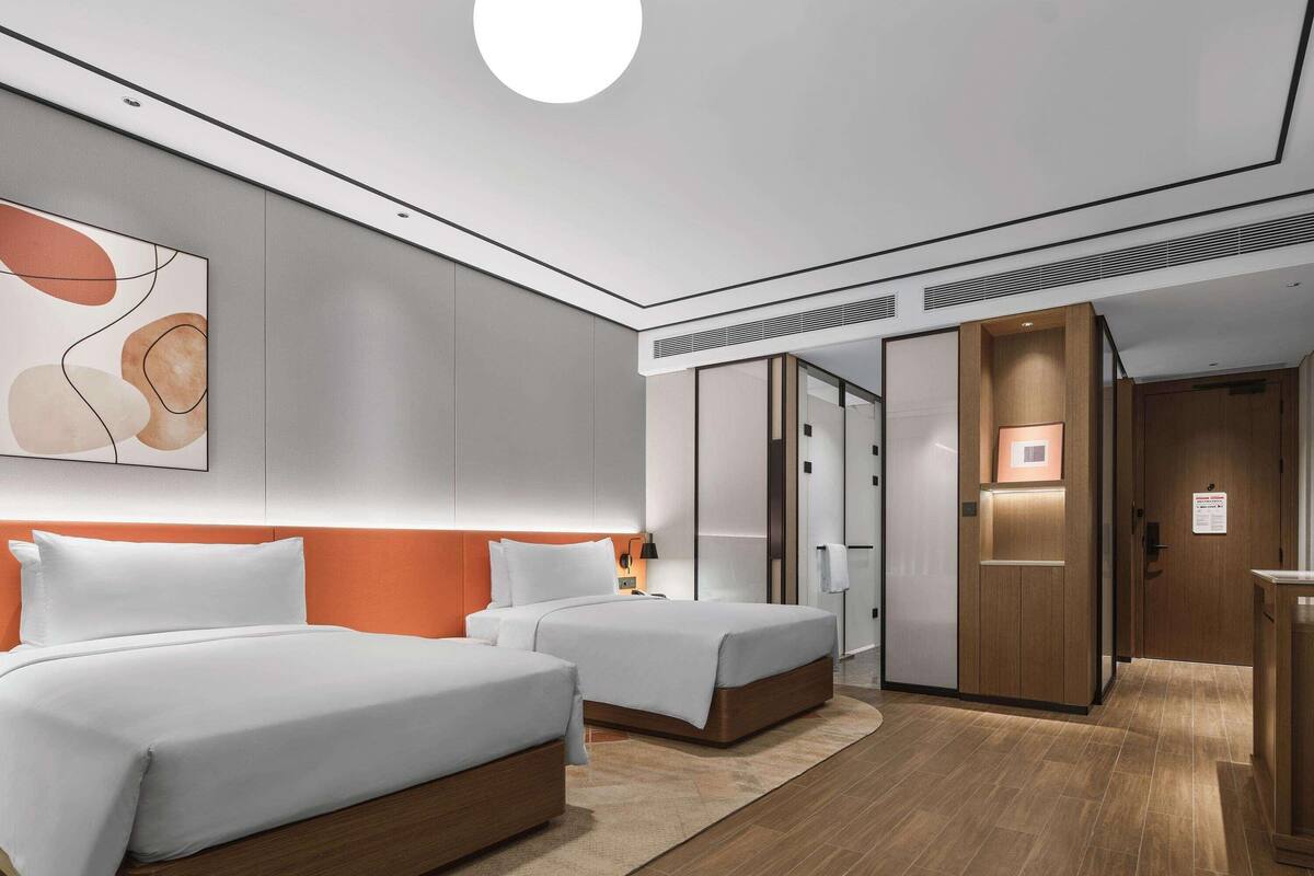 Une chambre d’hôtel moderne avec deux lits, une grande peinture abstraite, un luminaire rond au plafond et un plancher en bois.