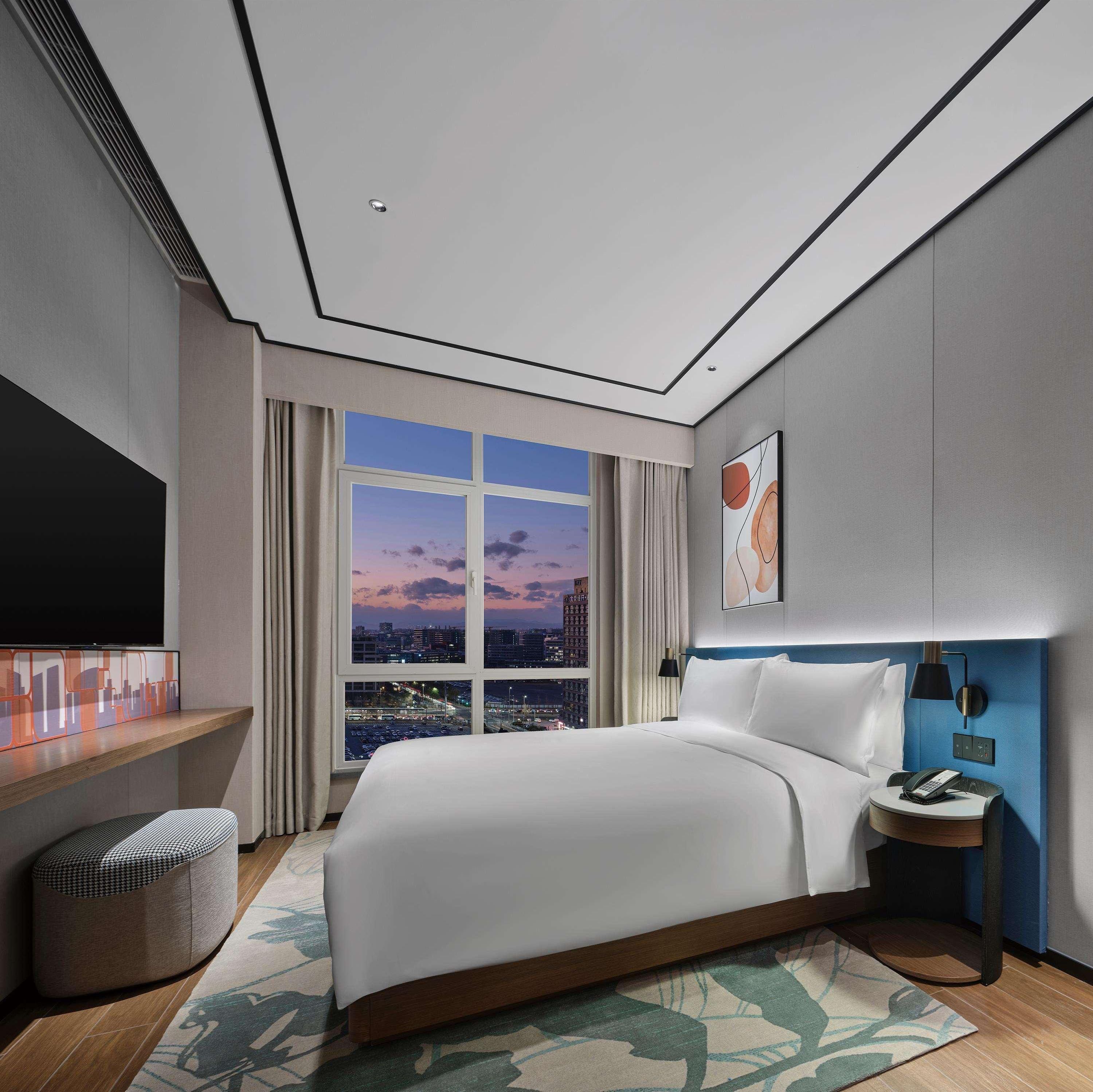 Quarto de hotel moderno com uma cama grande, televisão, escrivaninha e vista para a cidade ao entardecer.