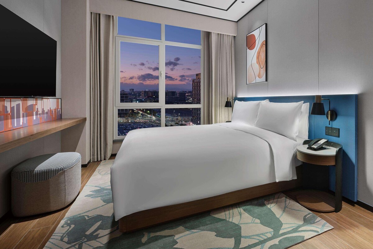 A modern hotel room with a large bed, a TV, a desk, and a view of the cityscape at dusk.