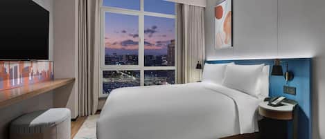 A modern hotel room with a large bed, a TV, a desk, and a view of the cityscape at dusk.