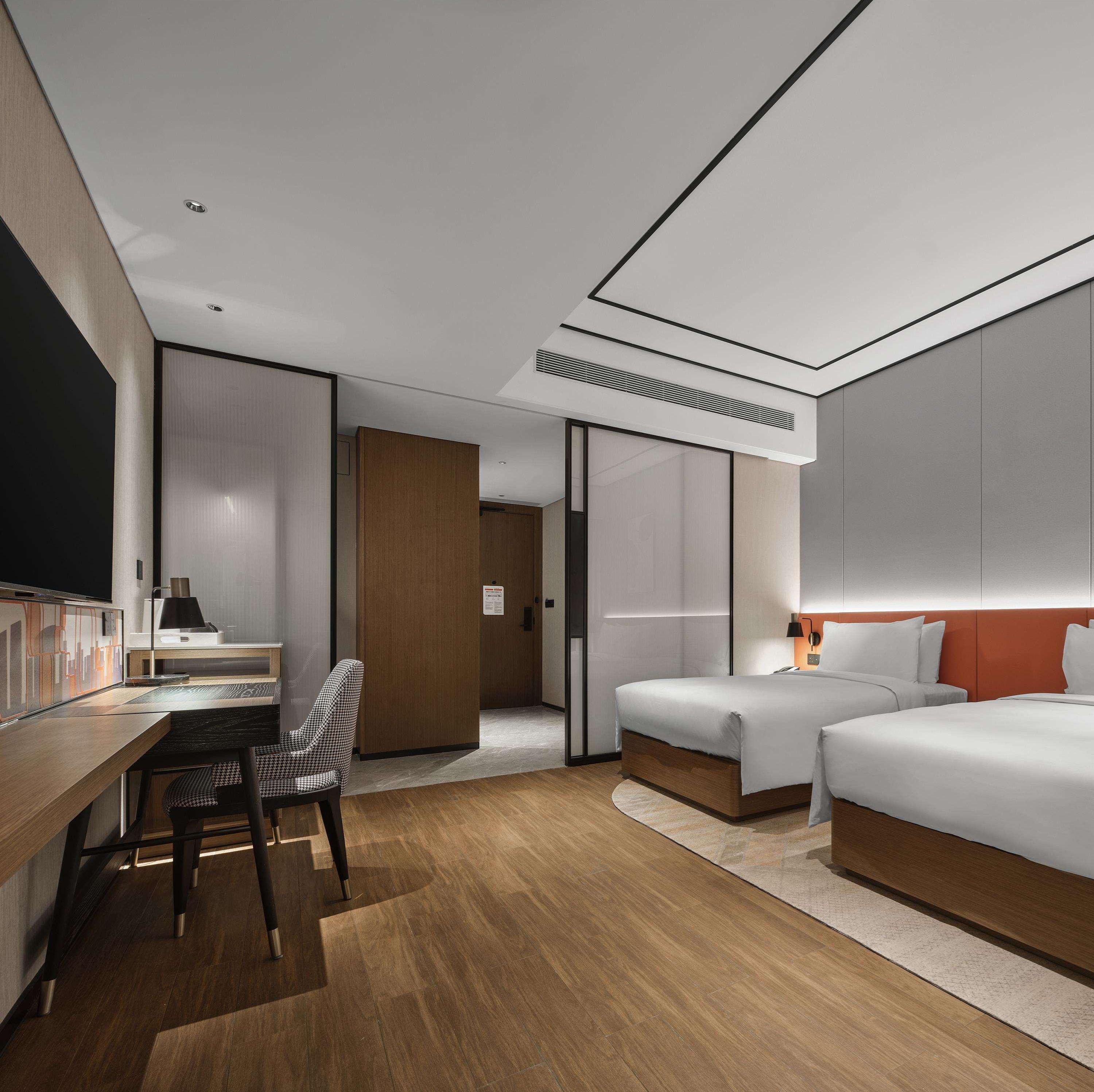 A modern hotel room with a wooden desk, two beds, a chair, and a large mirror.