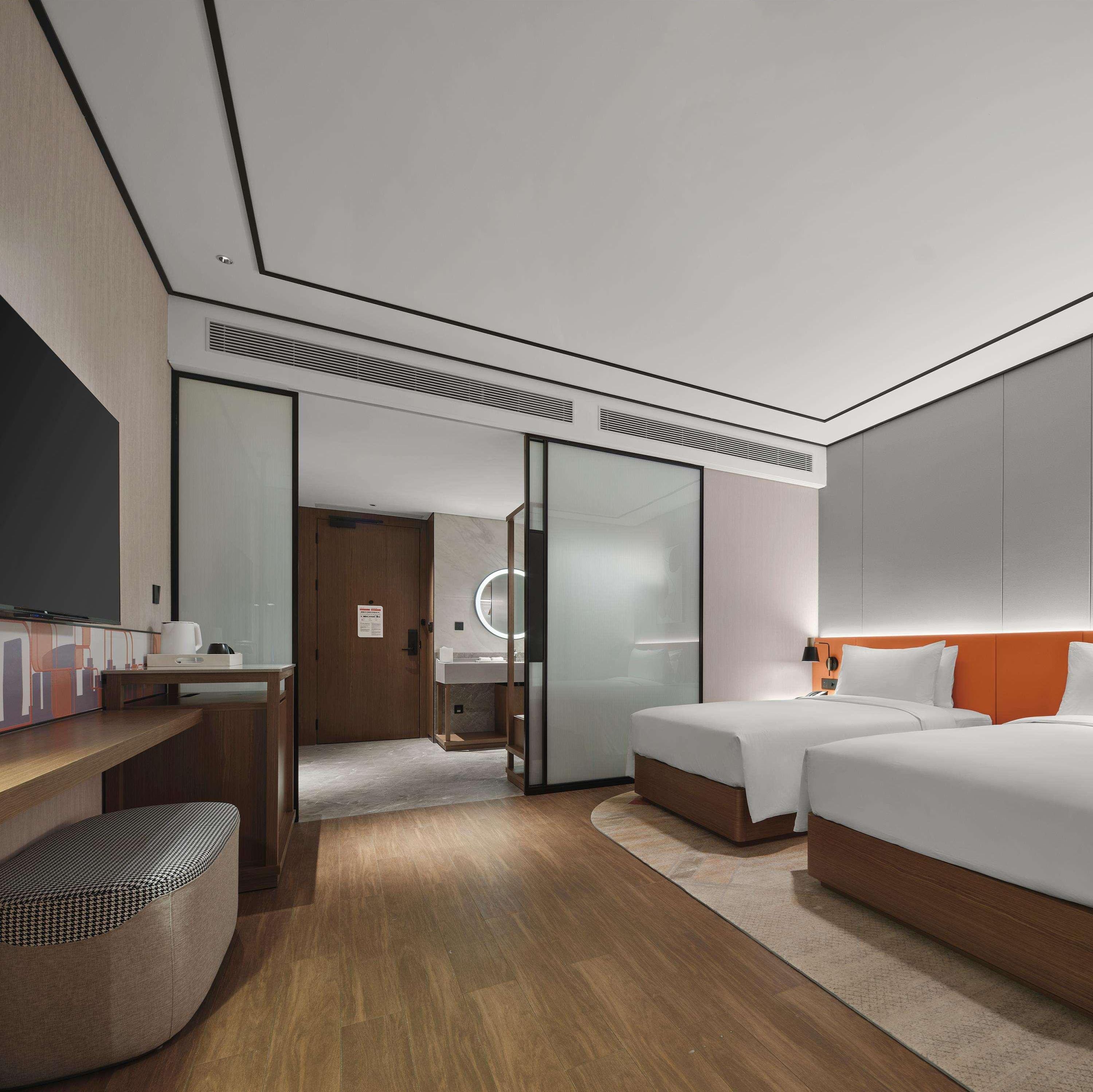 A modern hotel room with a wooden desk, a bed with white bedding, a large mirror, and a separate bathroom area.