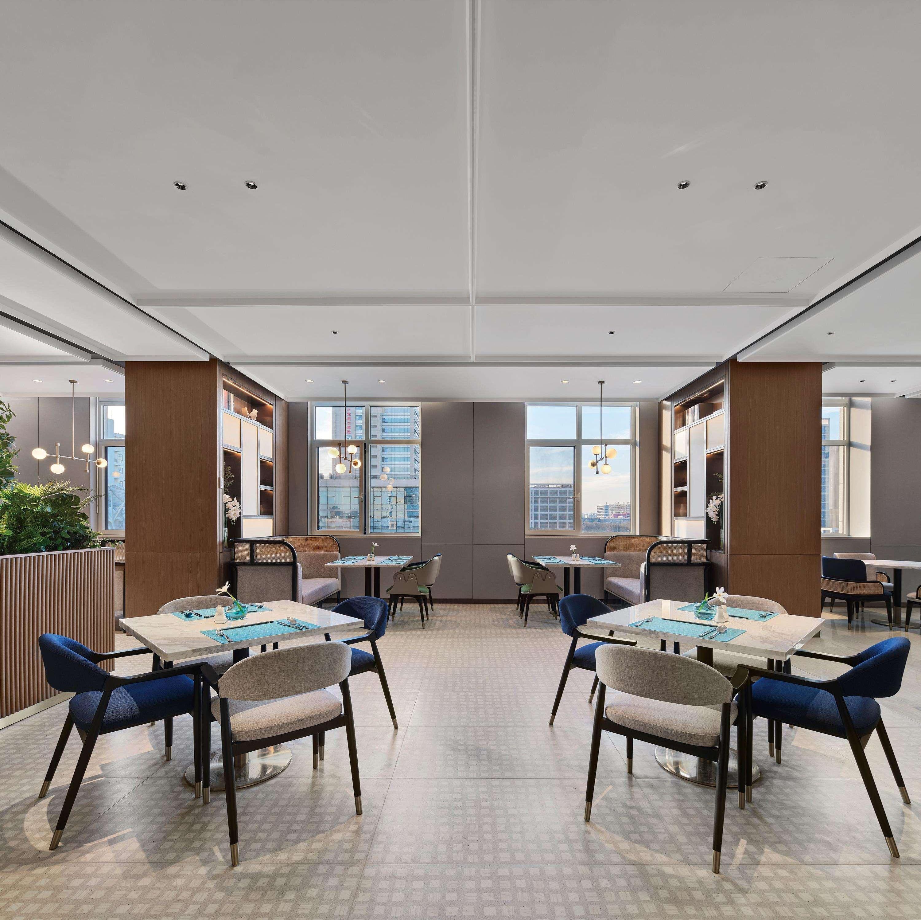 A modern dining area with multiple tables and chairs, large windows with city views, and a high ceiling with recessed lighting.