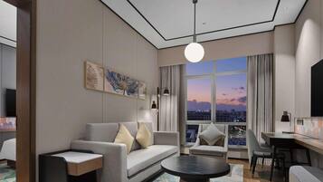 A modern hotel room with a grey sofa, a coffee table, and a city view through large windows.