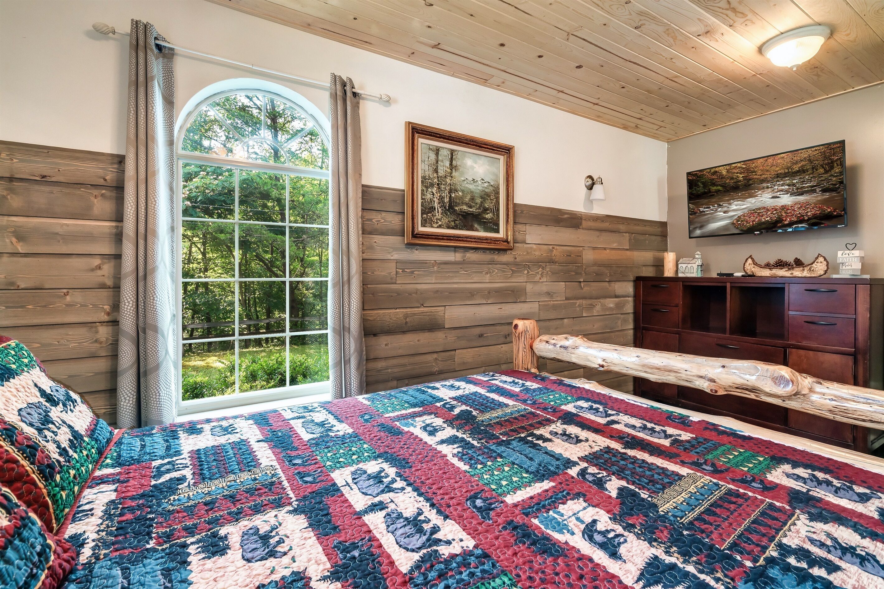 A cozy bedroom with a large window, wooden walls, a quilted bedspread, and a wooden headboard.