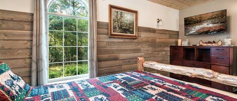A cozy bedroom with a large window, wooden walls, a quilted bedspread, and a wooden headboard.
