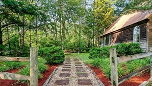 A wooden cabin with a gravel pathway leading to it, surrounded by dense greenery and a wooden fence.