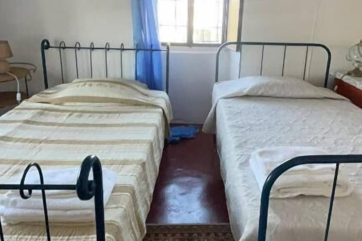 Quarto pequeno e simples com duas camas de metal, uma janela com cortinas azuis e um tapete estampado no chão.
