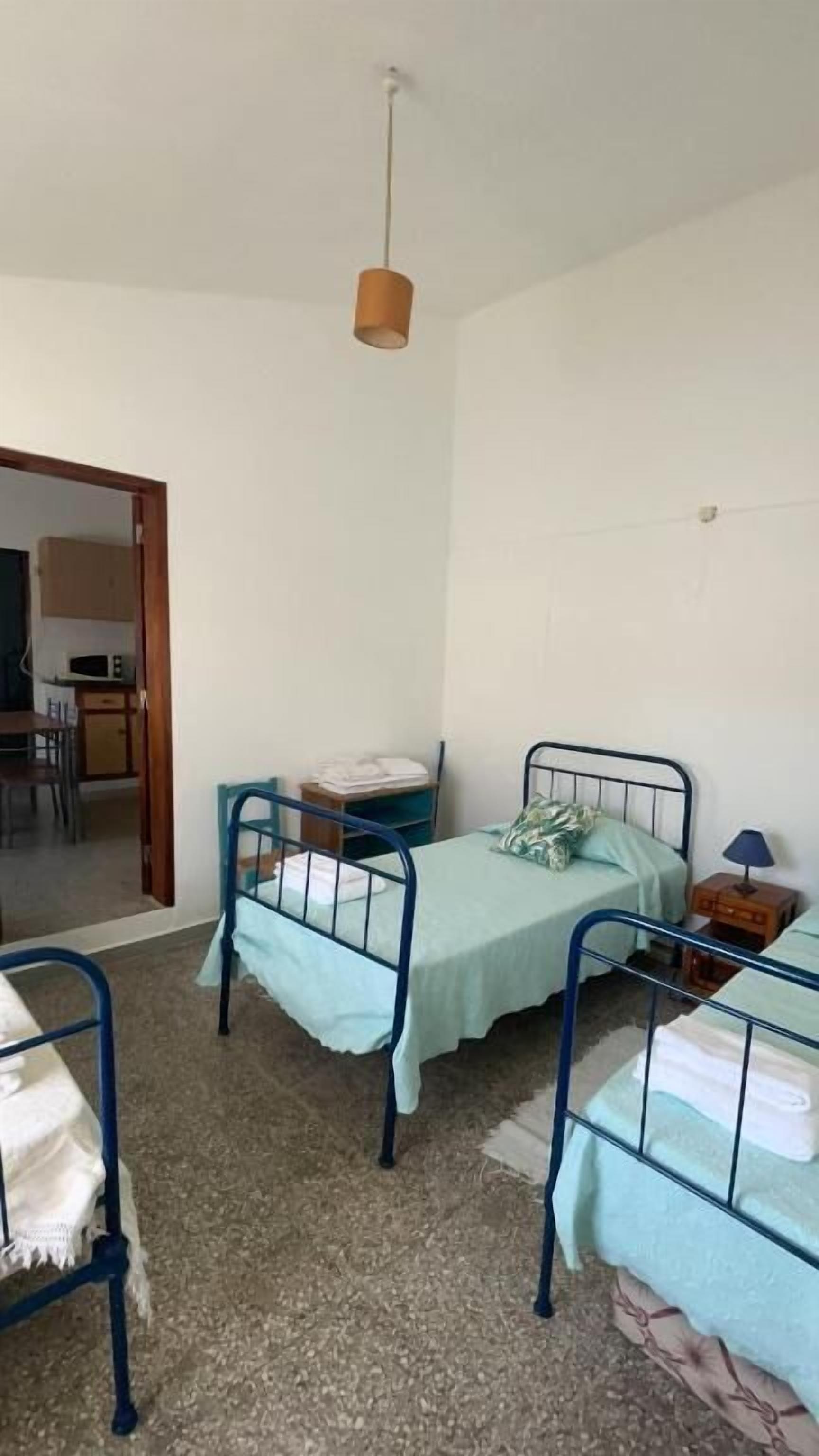 Una habitación pequeña y sencilla con dos literas metálicas, una silla, un escritorio y una lámpara colgante.