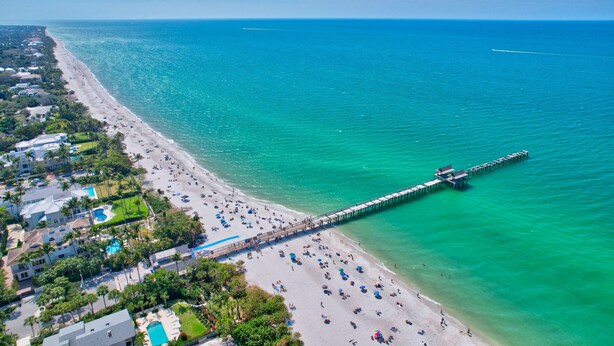 An aerial view of a beach with a pier extending into the ocean, surrounded by buildings and greenery. - Olde Naples beautiful Condo Steps to Pier, Beach, Sunsets and restaurants!  (Naples)
