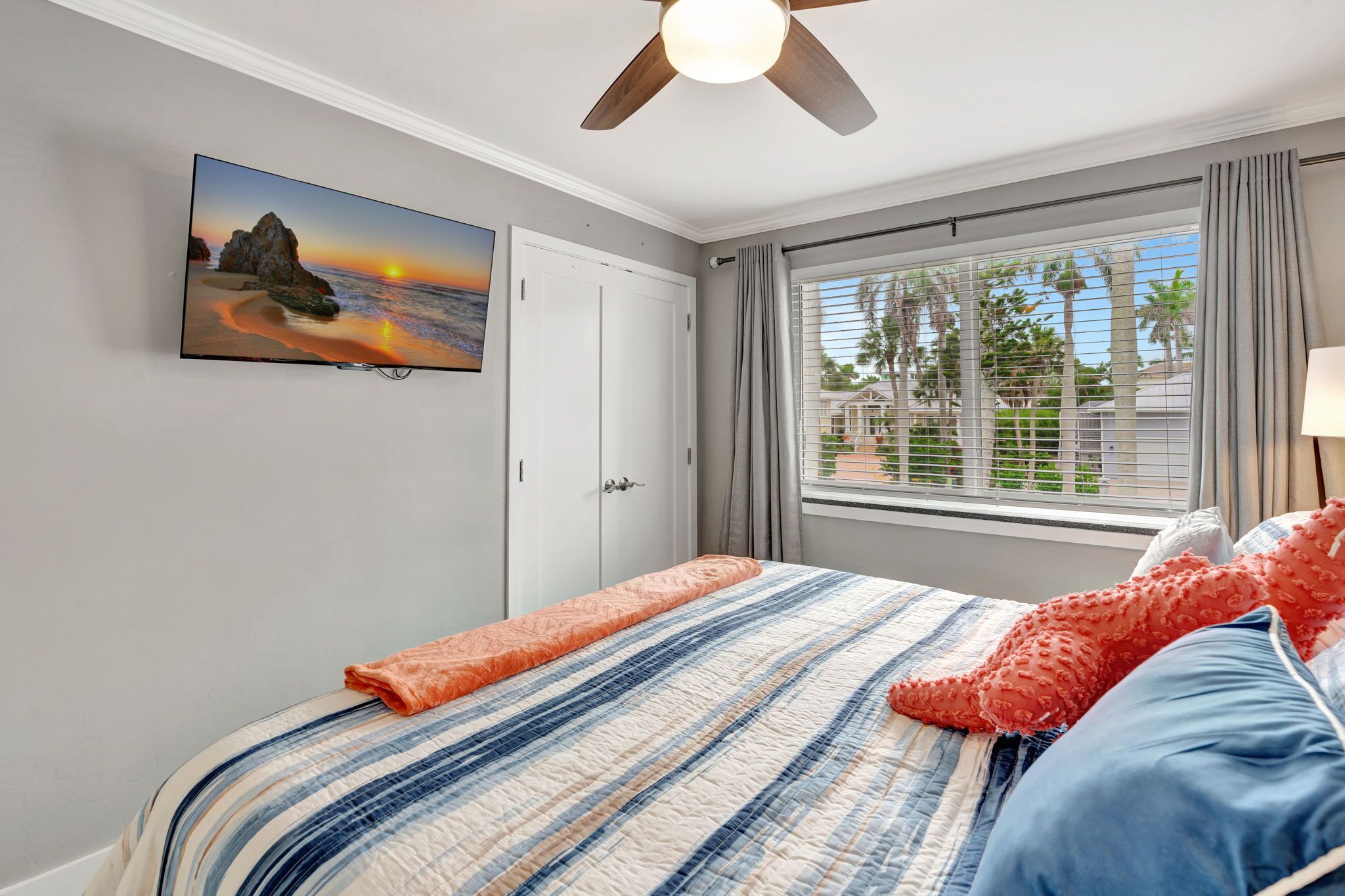 Een slaapkamer met een bed, een plafondventilator, een raam met gordijnen en een aan de muur bevestigde televisie waarop een zonsondergang te zien is.