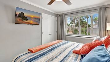 Een slaapkamer met een bed, een plafondventilator, een raam met gordijnen en een aan de muur bevestigde televisie waarop een zonsondergang te zien is.