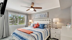 A bedroom with a bed, a ceiling fan, a window with curtains, a nightstand with a lamp, and a painting on the wall. - Olde Naples beautiful Condo Steps to Pier, Beach, Sunsets and restaurants!  (Naples)