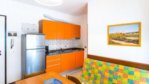 Une cuisine de taille réduite avec des placards orange, un réfrigérateur et une cuisinière. On y trouve une table en bois, un panier bleu et un banc matelassé aux couleurs vives. Un tableau représentant un champ de tournesols est accroché au mur.