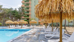 Un espace piscine doté de chaises longues et de parasols en chaume, entouré d’une clôture et d’arbres.