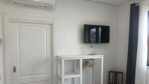 Una habitación con puerta blanca, estantería blanca, televisor montado en la pared, una silla y una cama con cubrecama blanco.