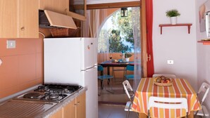 A compact kitchen with a gas stove, a white refrigerator, and wooden cabinets. There is a dining area with a table covered by a striped tablecloth and chairs. A small potted plant is on a shelf above the table. - [Dimora dell'Arco] Seaside Apartment with Stunning Views (Orosei)