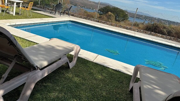 Una piscina con dos sillas de descanso y una mesa, rodeada por una cerca de alambre y vistas a un lago y colinas.