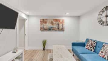 A modern living room with a blue sofa, a white coffee table, a painting on the wall, and a clock.