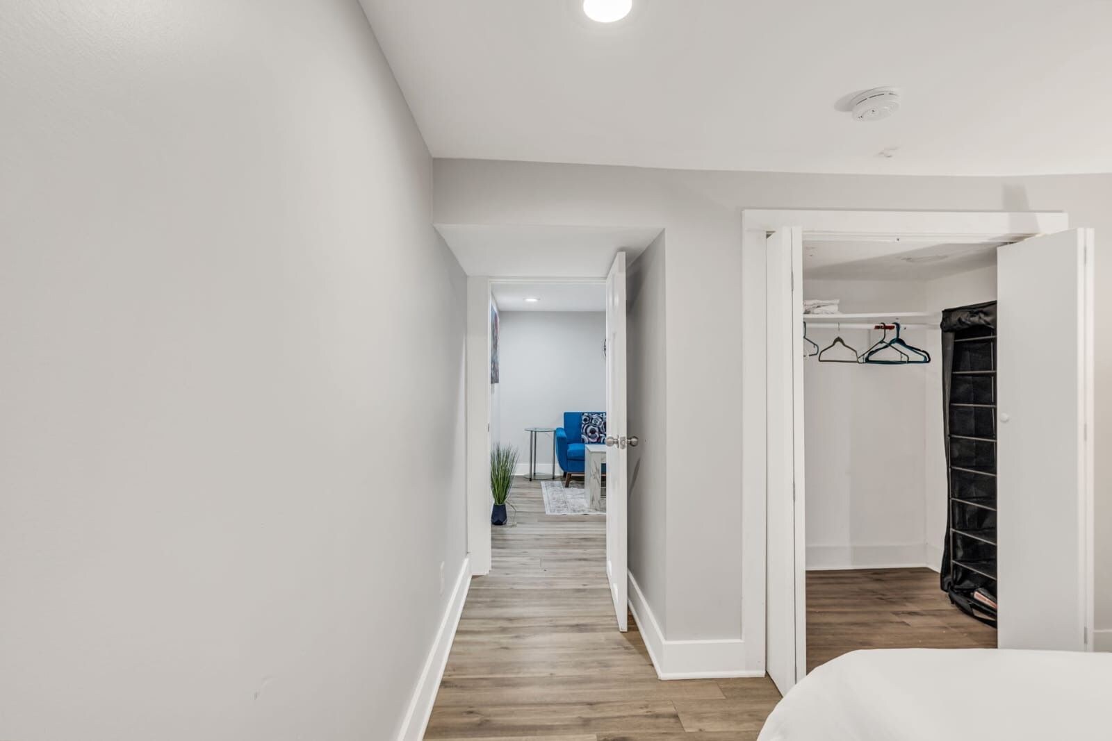 Un pasillo con piso de madera, una puerta blanca y una habitación con cama y un mueble de almacenaje negro.