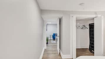 A hallway with a wooden floor, a white door, and a room with a bed and a black storage unit.