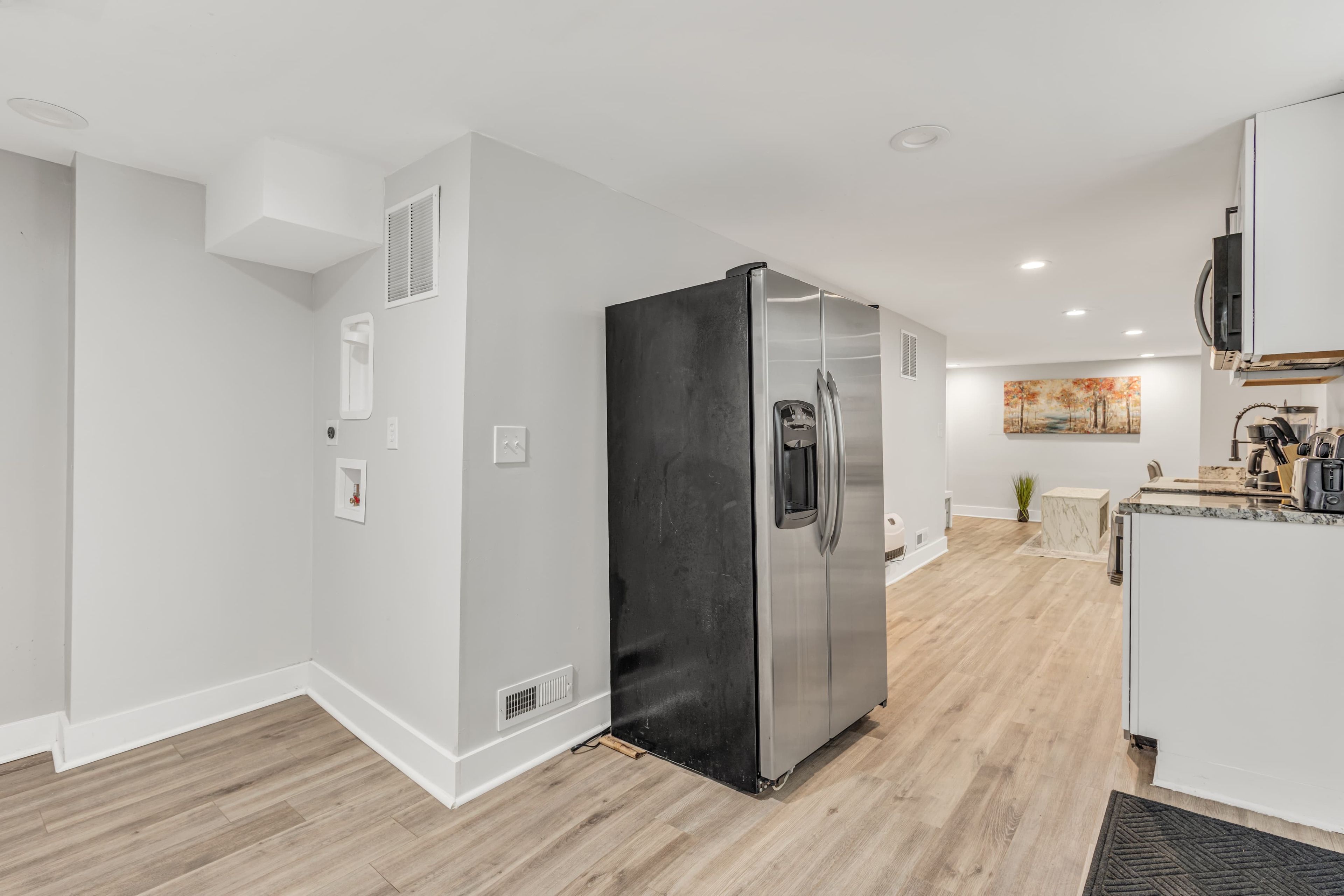 A modern kitchen with a stainless steel refrigerator, wooden flooring, white walls, and recessed ceiling lighting.
