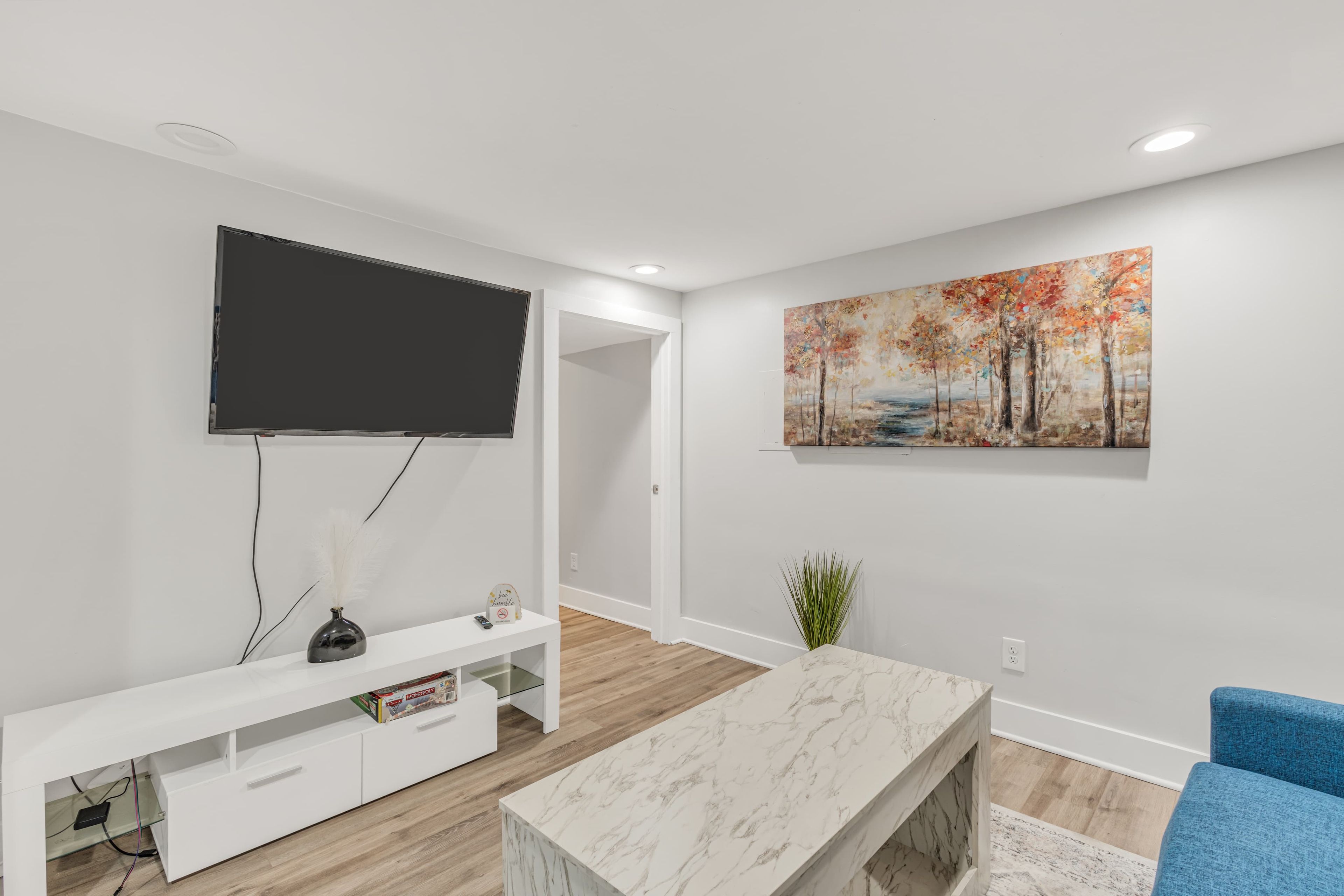 Ein modernes Wohnzimmer mit Flachbildfernseher, Marmortisch, blauem Sessel, weißem Lowboard und einem Gemälde an der Wand.