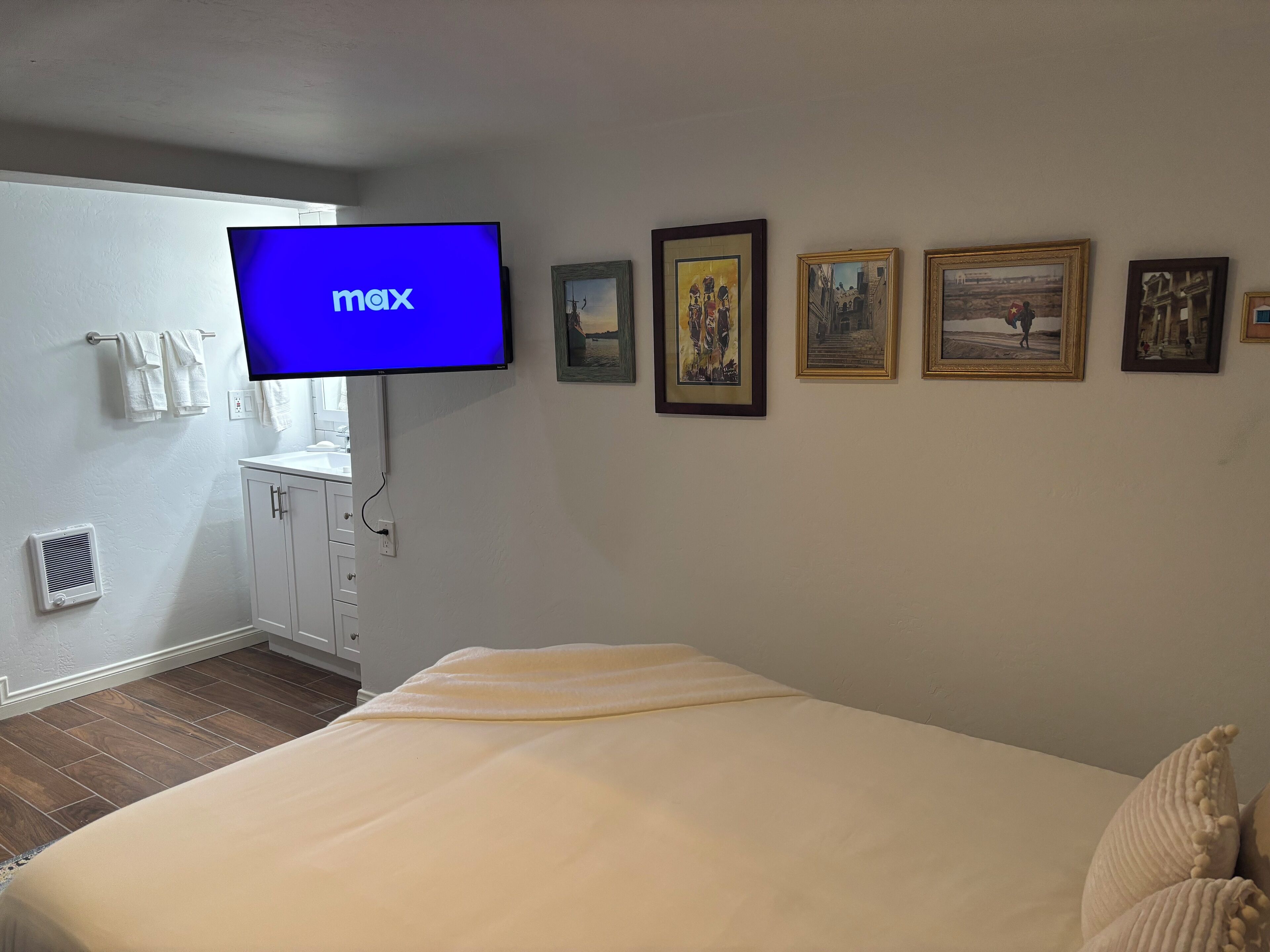 A hotel room with a bed, a TV mounted on the wall, a bathroom with a sink and towel rack, and several framed pictures on the wall.