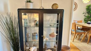 A glass-fronted cabinet with decorative items, a potted plant, and a dining area with wooden furniture. - The Beachcomber Oceanside Getaway! NEW 2025 (Crescent City)