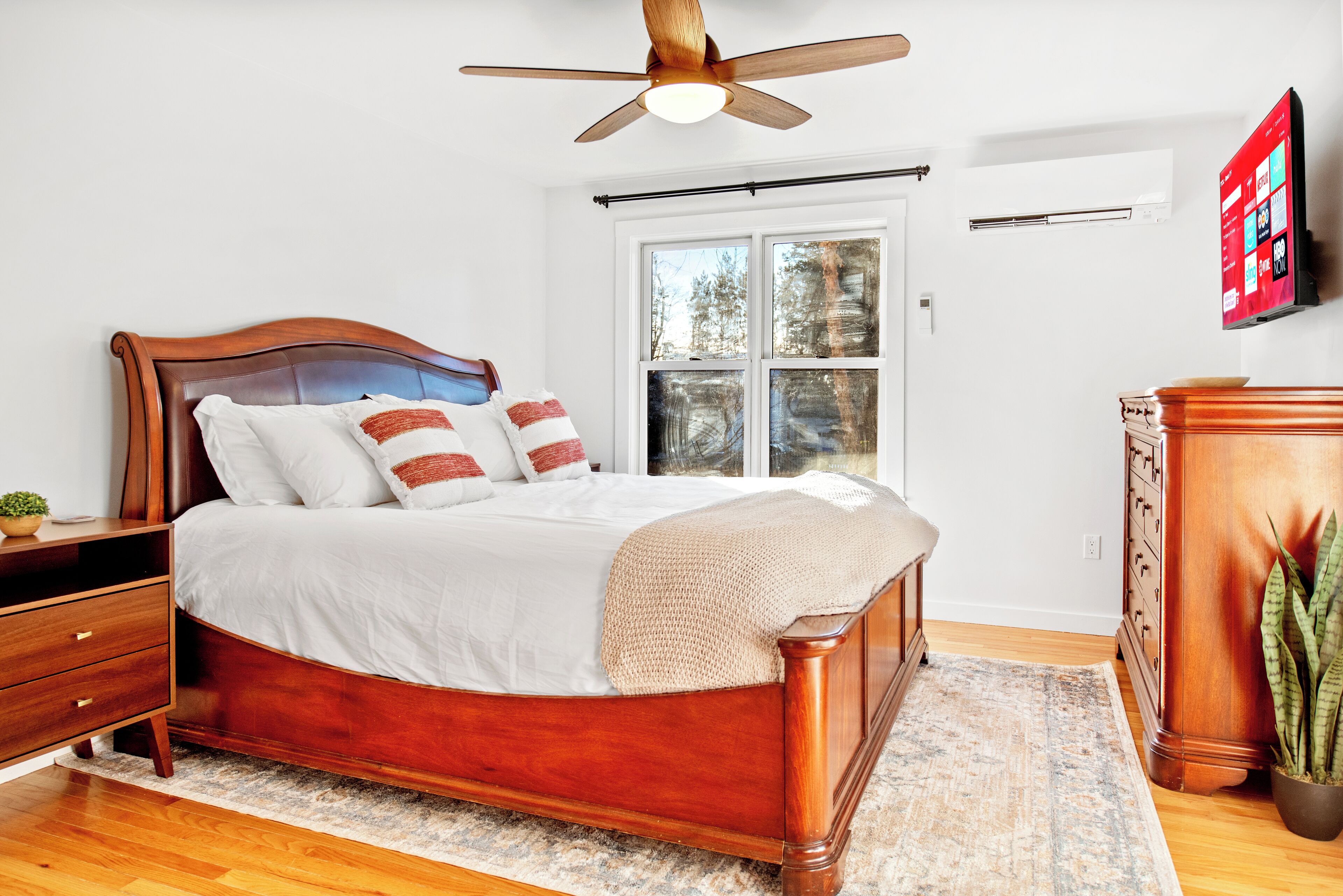 A bedroom with a wooden bed, a ceiling fan, a window with blinds, a dresser, and a television on the wall.