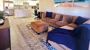 Un salon moderne avec un canapé beige, une couverture bleue et un tapis à motifs. On aperçoit au fond une cuisine avec des meubles blancs et un réfrigérateur.