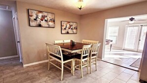 Un coin repas avec une table et des chaises en bois, deux tableaux abstraits accrochés au mur, une bouche d’aération au plafond et une porte donnant sur une autre pièce.