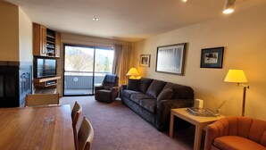 A living room with a fireplace, a television, a sofa, a dining table with chairs, and a balcony with a view of trees. - Cozy condo, next to the ski slopes with 2 private balconies (Whitefish)