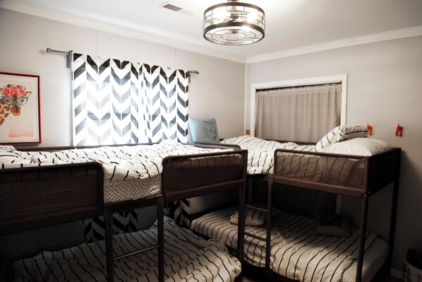 A dormitory room with bunk beds, a bed with a patterned bedspread, a framed picture on the wall, and a window with curtains.