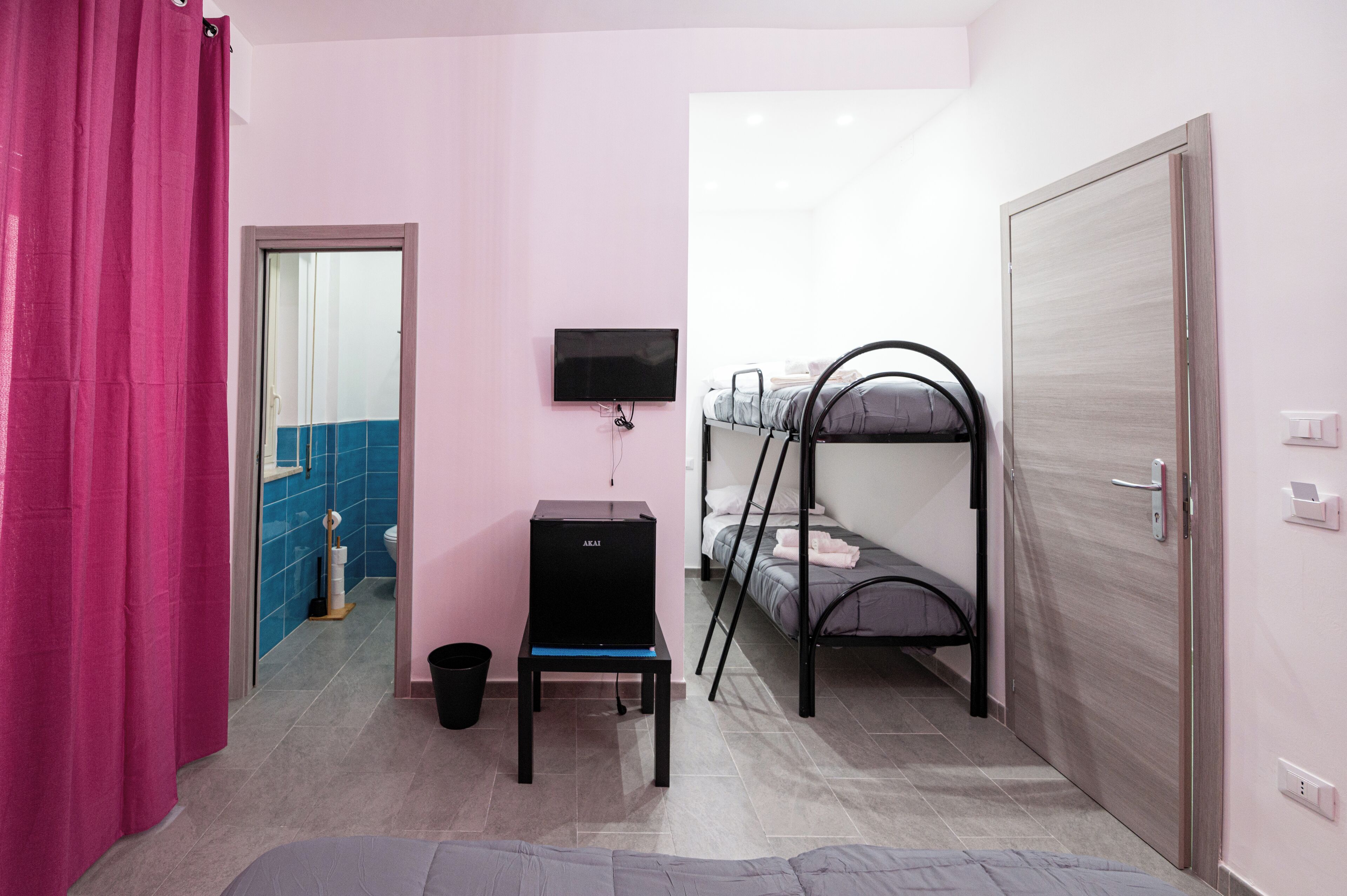 Une petite pièce avec un lit superposé, un bureau avec un ordinateur, une télévision et un rideau rose.
