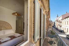 A hotel room with a bed, a hanging light fixture, a balcony with chairs, and a view of the street.