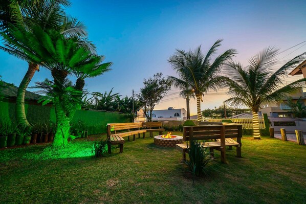 An outdoor seating area with wooden benches and a fire pit, surrounded by palm trees and a green fence. - AP 08A Ala Morrinhos Residencial Solavir (Bombinhas)