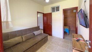 A compact living space with a brown sofa, a wooden door, a small table with a TV, and a tiled floor. - AP 08A Ala Morrinhos Residencial Solavir (Bombinhas)