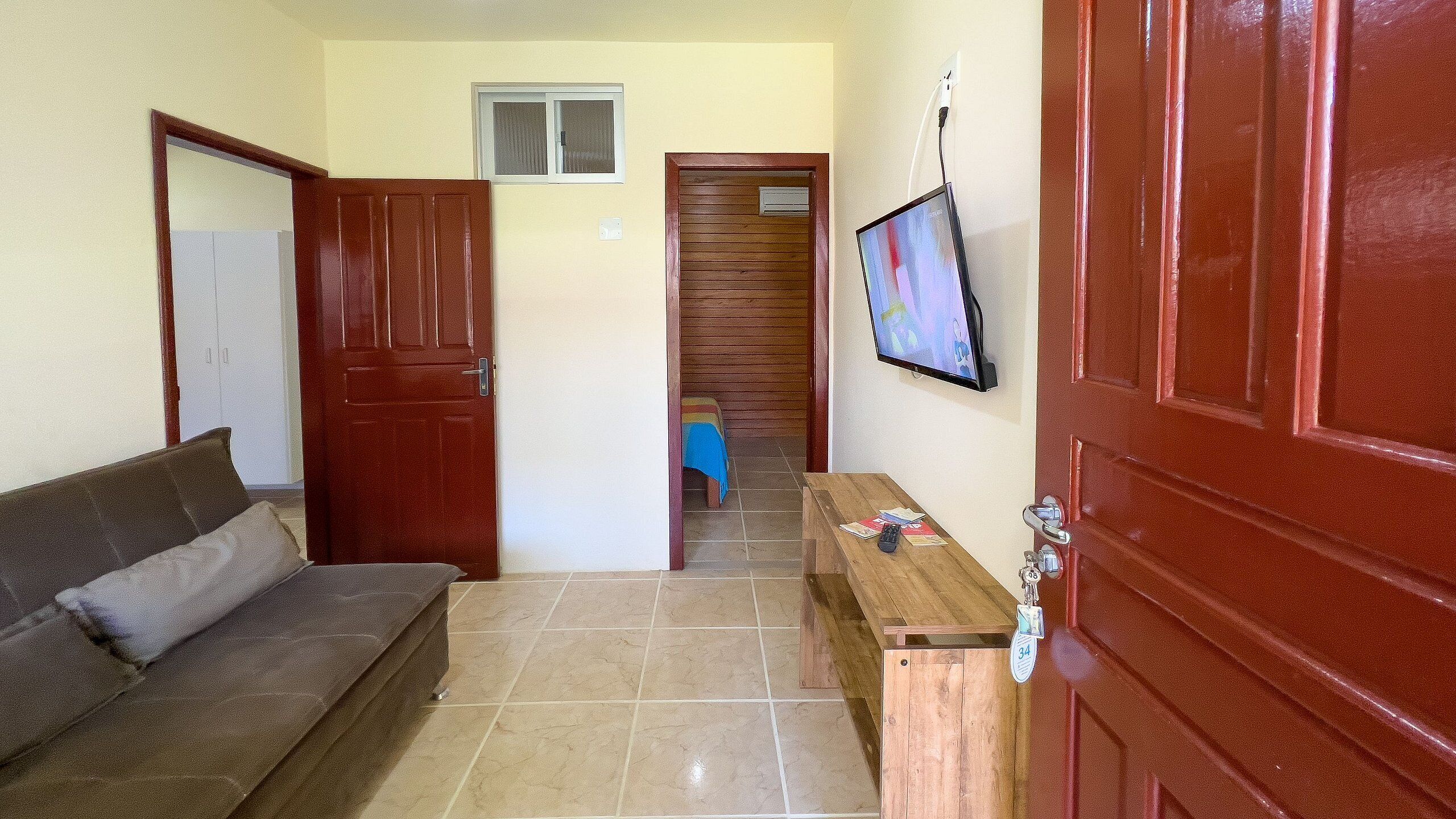 Um corredor com uma porta de madeira, um sofá marrom, uma mesa pequena com objetos e uma televisão na parede.
