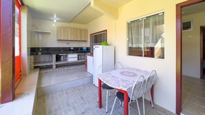 A compact kitchen with a refrigerator, a sink, and a window with curtains. There is a dining table with chairs and a tiled floor.