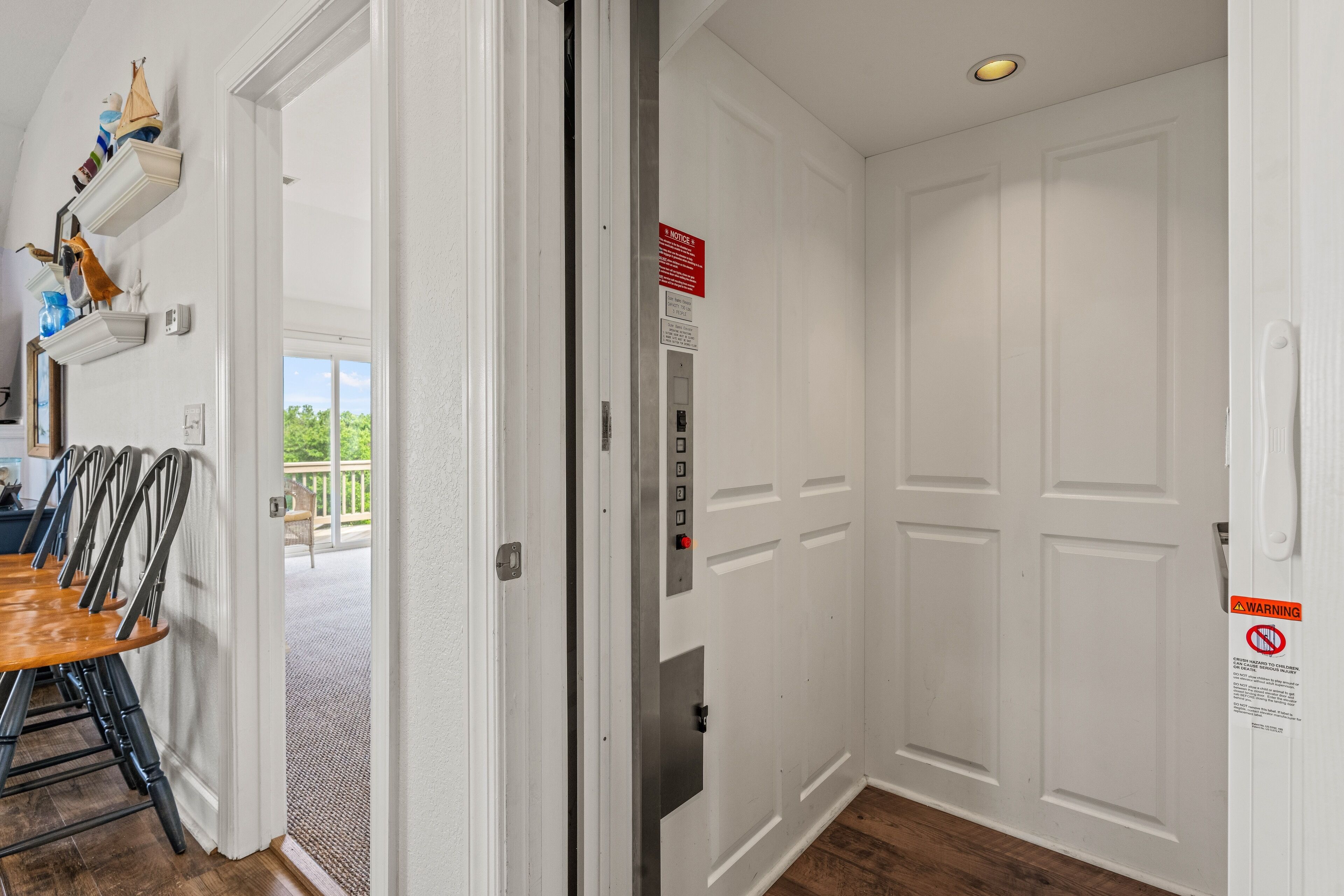 A hotel room with a wooden dining table, chairs, a white door with a warning sign, and a view of the outdoors.