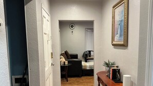 Un pasillo con suelo de madera, una puerta, una mesita con un florero, un reloj y un cuadro en la pared.