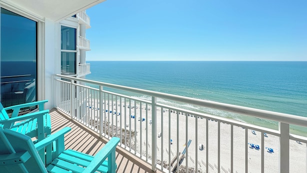A balcony with turquoise chairs, a view of the ocean, and a sandy beach with umbrellas.