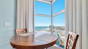 A dining area with a wooden table and chairs, large windows with white curtains, and a view of the beach and cityscape.
