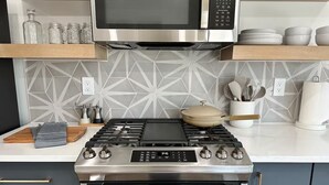 A modern kitchen with a stainless steel stove, oven, and microwave. There are wooden shelves above the stove holding dishes and jars. A cutting board and a cloth are on the counter. - Bright and Modern Studio Guesthouse - near RiNo! (Denver)