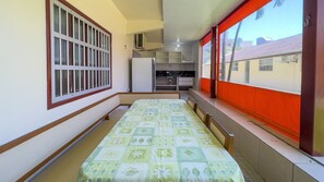 A narrow room with a bed, a window with bars, a kitchenette, and a view of buildings outside.
